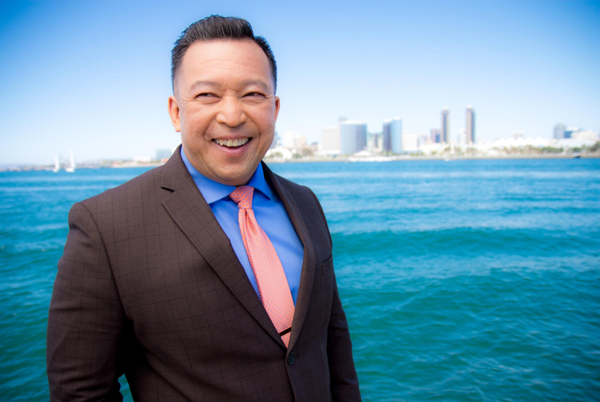 Smiling executive in a suit standing by the ocean, with a city skyline in the background — captured in natural light for a bold and confident portrait.