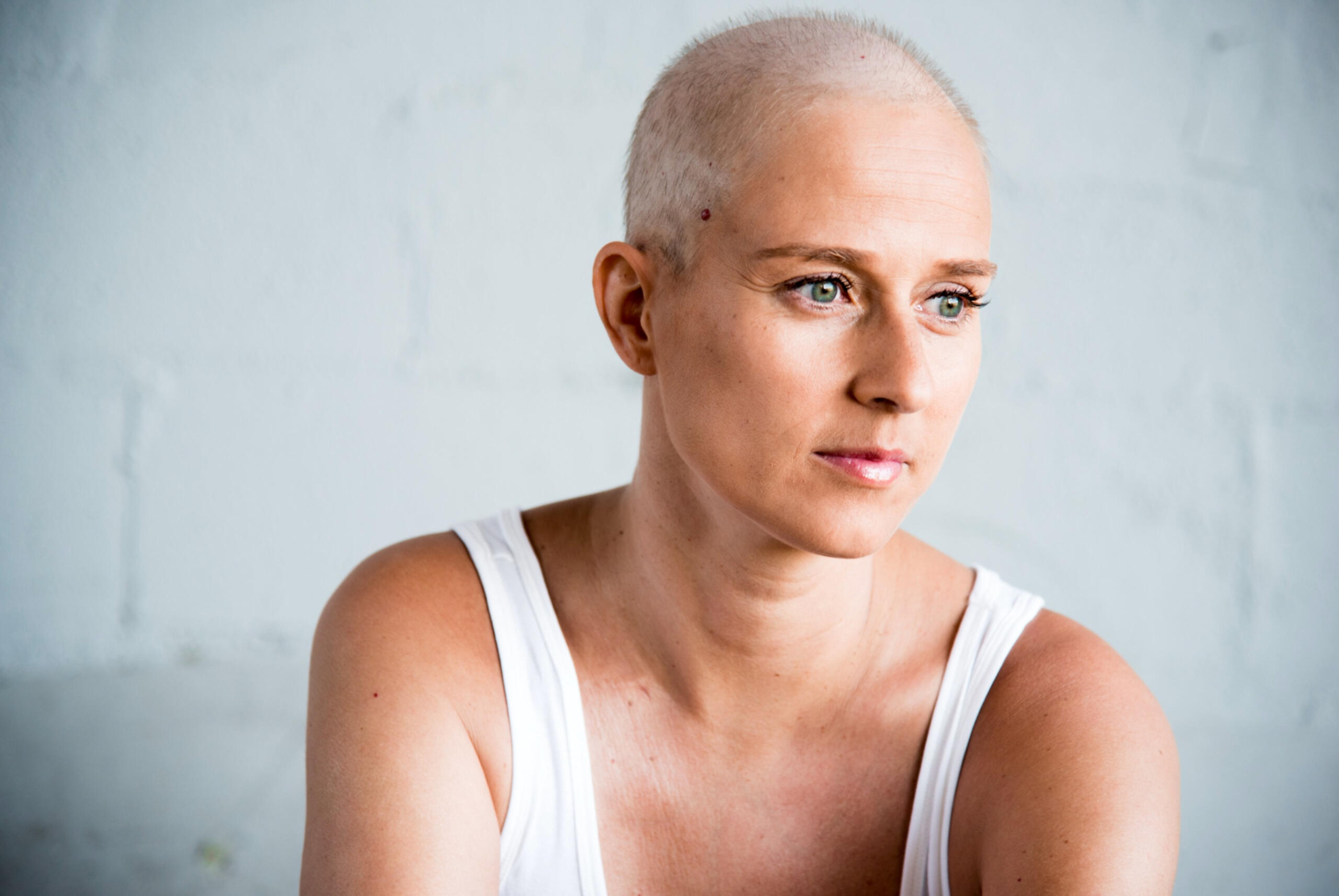Woman with shaved head gazing softly off-camera in a minimalist white tank top — an intimate, unguarded portrait bathed in natural light.